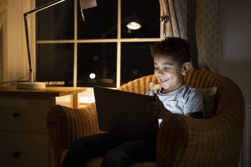 A close-up shot of a boy using a digital tablet while sitting on a chair. He is laughing while playing on the tablet in a low lit room.